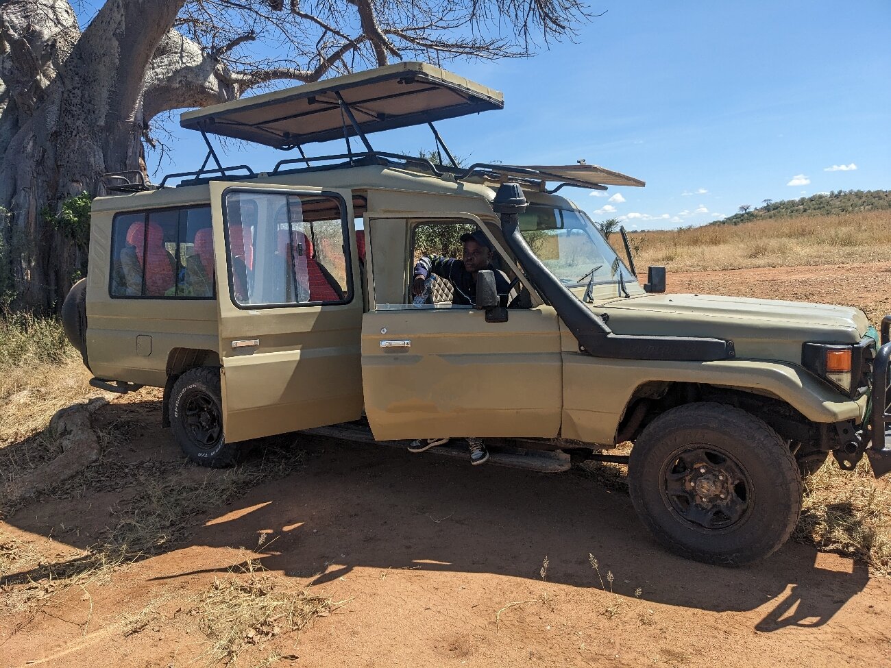A safari vehicle and its driver-guide parked in the shade of a baobab in Ruaha